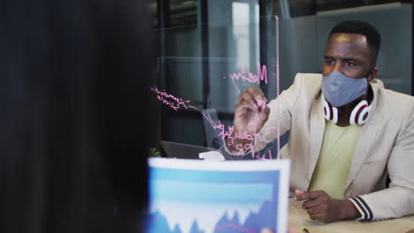 african american man wearing face mask writing on glass board at modern office