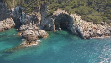 Cala-llonga-drone-aerial-view-revealing-tree-covered-cliffs,-blue-ocean,-sandy-beach-and-beautiful-resorts-on-the-Balearic-Islands-of-Ibiza,-Spain