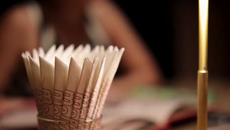 a hand reaches for a decorative cup of incense sticks on a table, with a blurred figure and open book in warm, soft lighting