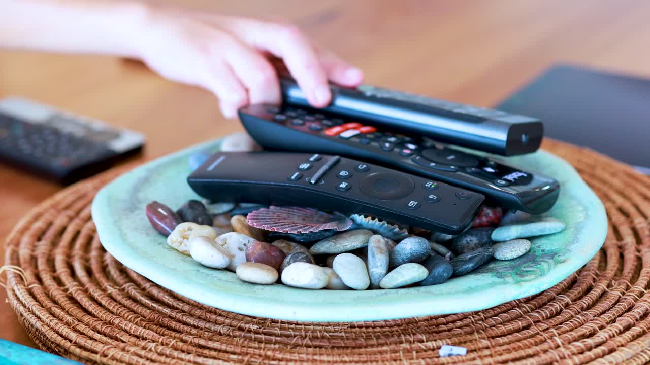 A person selects a remote control from a decorative plate filled with stones and shells on a wooden table, under bright natural lighting with steady camera framing