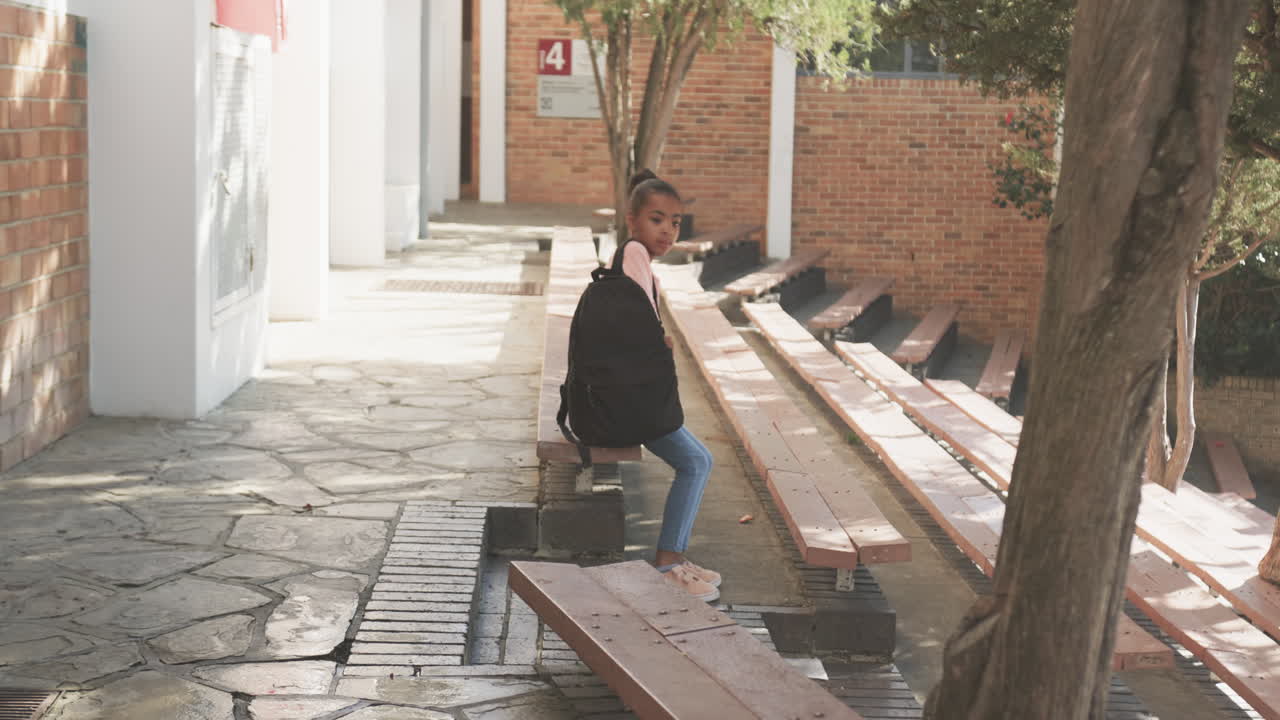 Sitting on bench, African American girl with backpack waiting outside school building