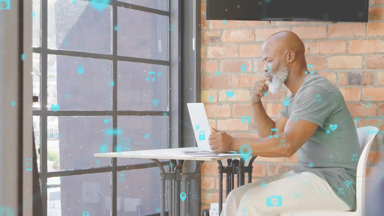 Mature man sitting at white table holding laptop while using hologram tech icons in tech office