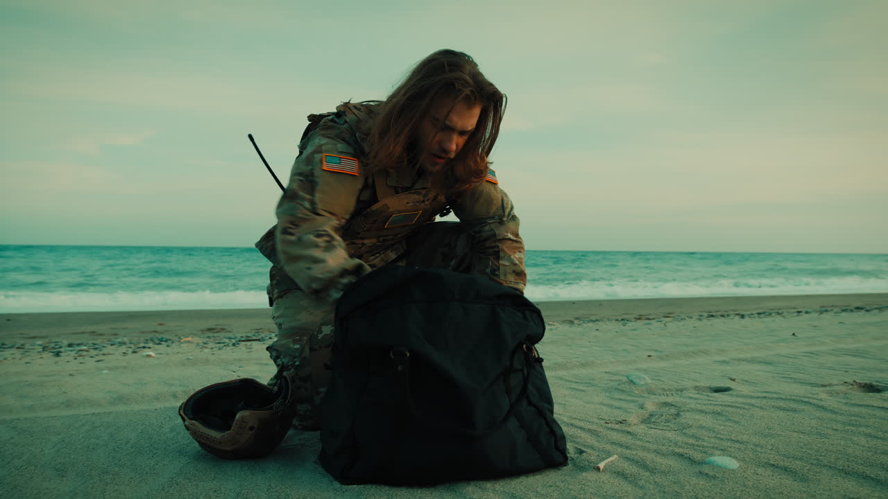 Charmante Amerikaanse soldaat met lang haar en rugzak op het strand