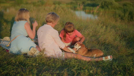 family enjoying outdoor picnic in field with playful dog, woman holds back dog gently while dog jumps up excitedly, little girl smiles at dog while another woman watches