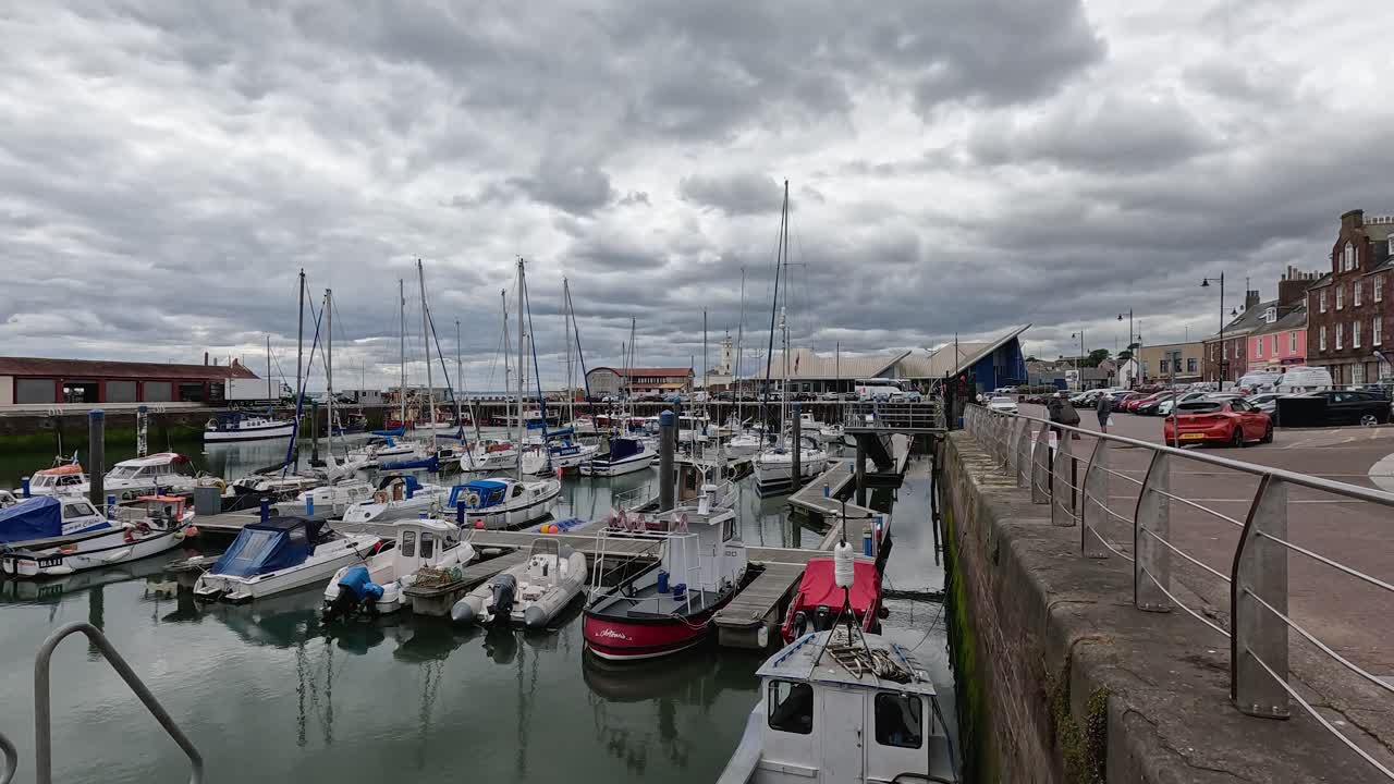 Static wide shot of marina boats, overcast sky, calm water, and waterfront buildings in Fife