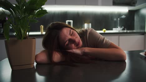 una chica morena con una camiseta beige descansa y se acuesta apoyándose en una mesa negra cerca de una planta de interior en un apartamento moderno en la cocina con cintas luminosas. tranquila chica feliz apoyándose en la mesa cerca de la flor interior en casa por la noche