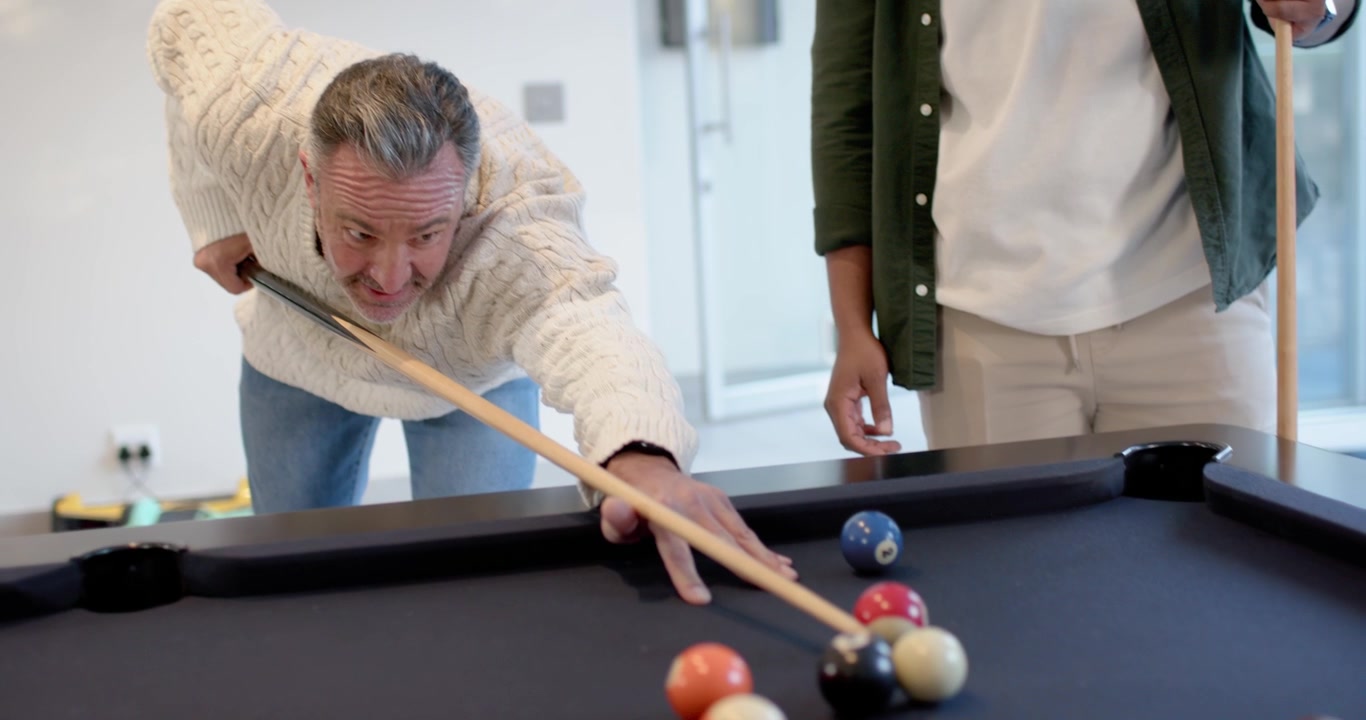 Diverse men watching senior man in sweater aiming and shooting cue ball, playing pool at home