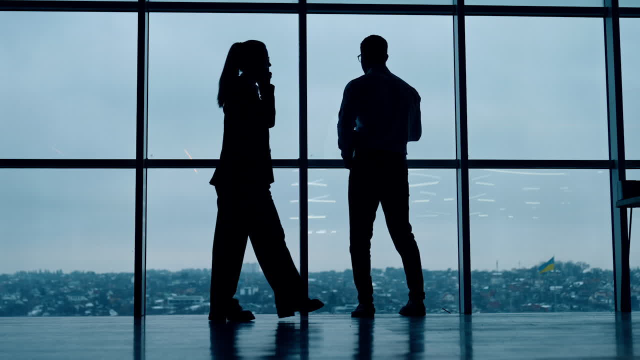 Silhouettes of business people working in the office with panoramic window. Man drinks coffee and woman has phone conversation.