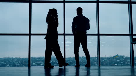 silhouettes of business people working in the office with panoramic window. man drinks coffee and woman has phone conversation.