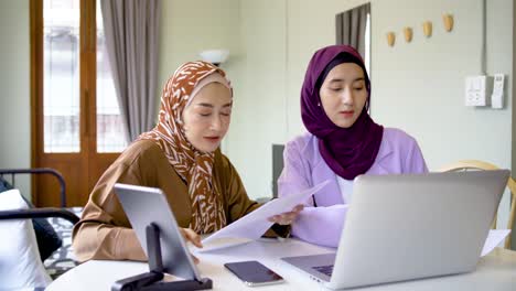 two asian muslim women wearing hijab working together with using laptop pc tablet and paper showing graphic chart in living room, talk about success working in an e-commerce business with a smile