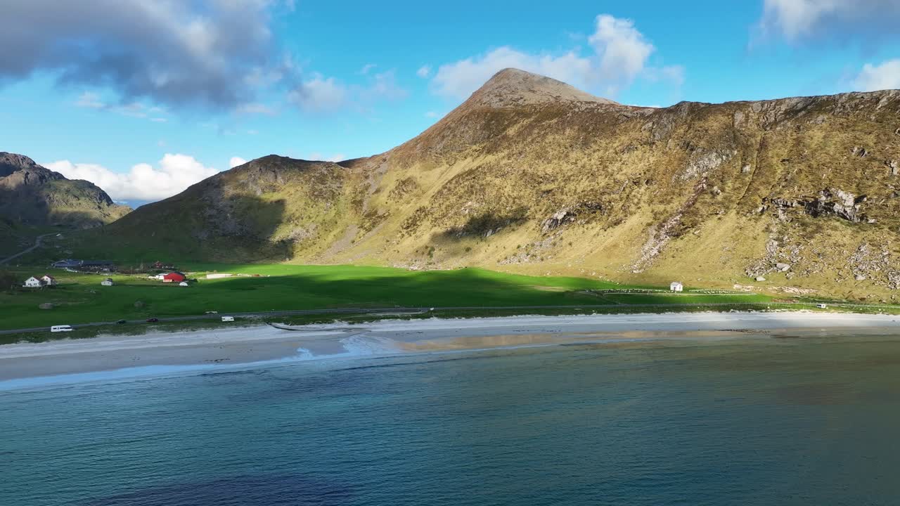 Calm aerial from sea towards Haukland beach with farms, green fields and mountains in evening sun