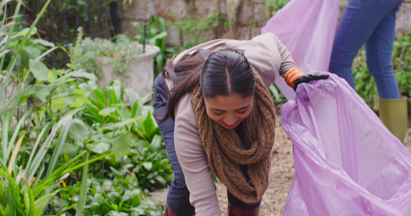 Due volontarie, una con una borsa rosa, l'altra che raccoglie i rifiuti con guanti arancioni, puliscono il giardino