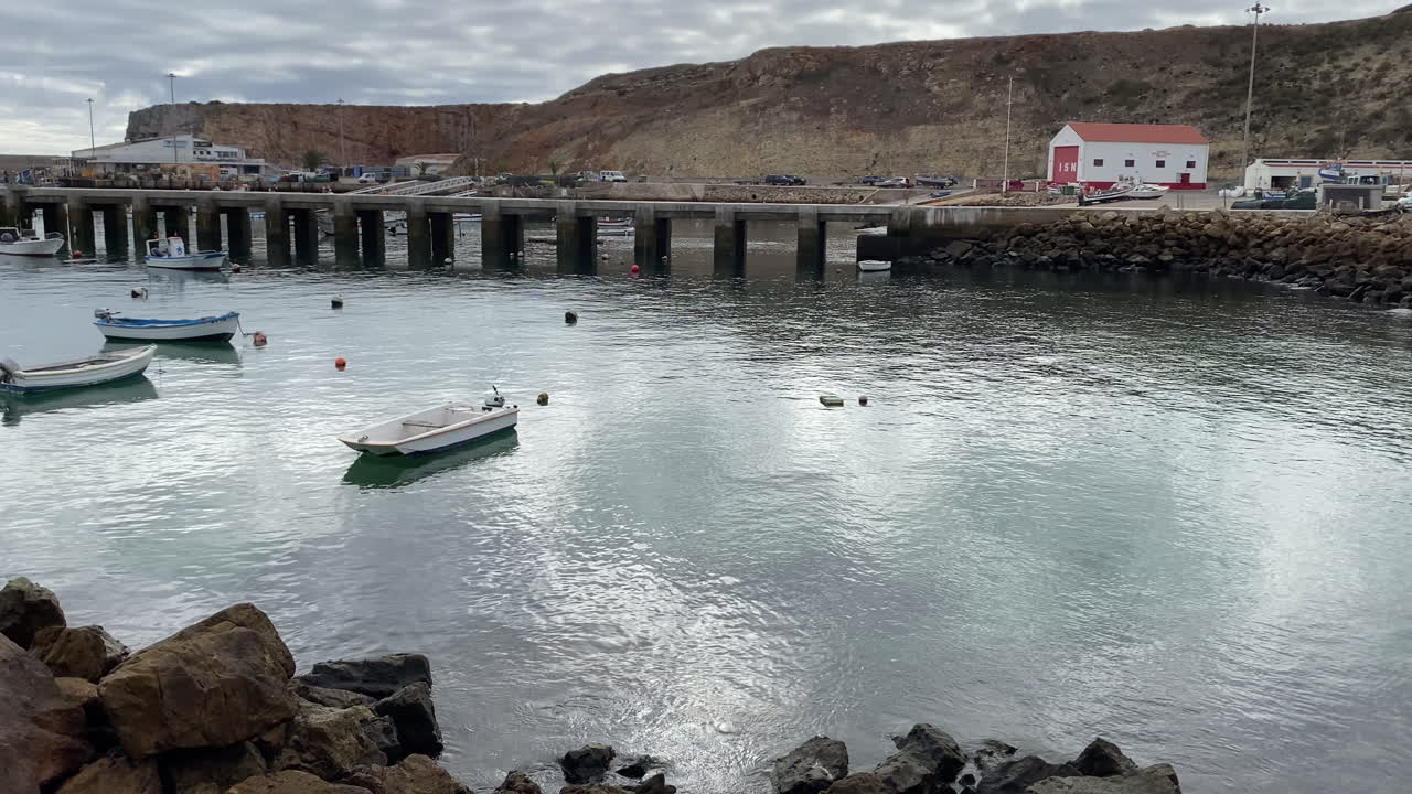 Fishing boats float calmly in Sagres harbor beneath golden cliffs and cloudy Atlantic skies travelling