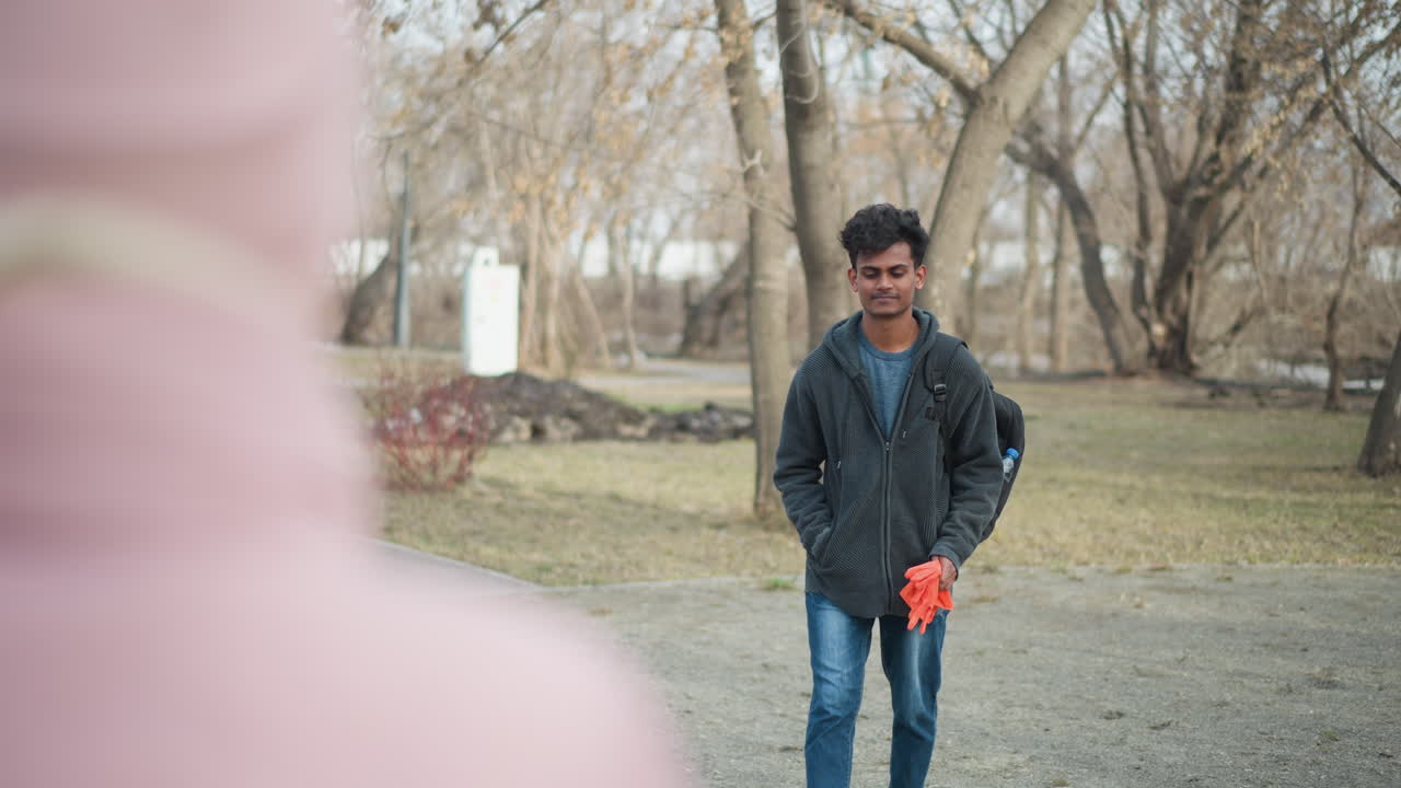 Young man with dark curly hair wearing gray hoodie and blue jeans standing outdoors in park holding orange gloves, carrying black backpack with water bottle, on early spring day