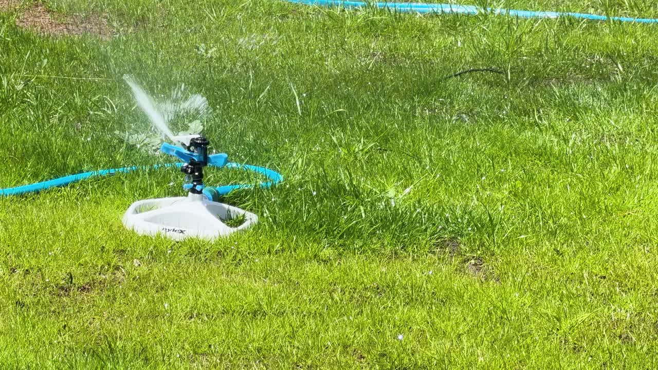 Slow motion rotating garden sprinkler waters bright green lawn under strong midday sunlight