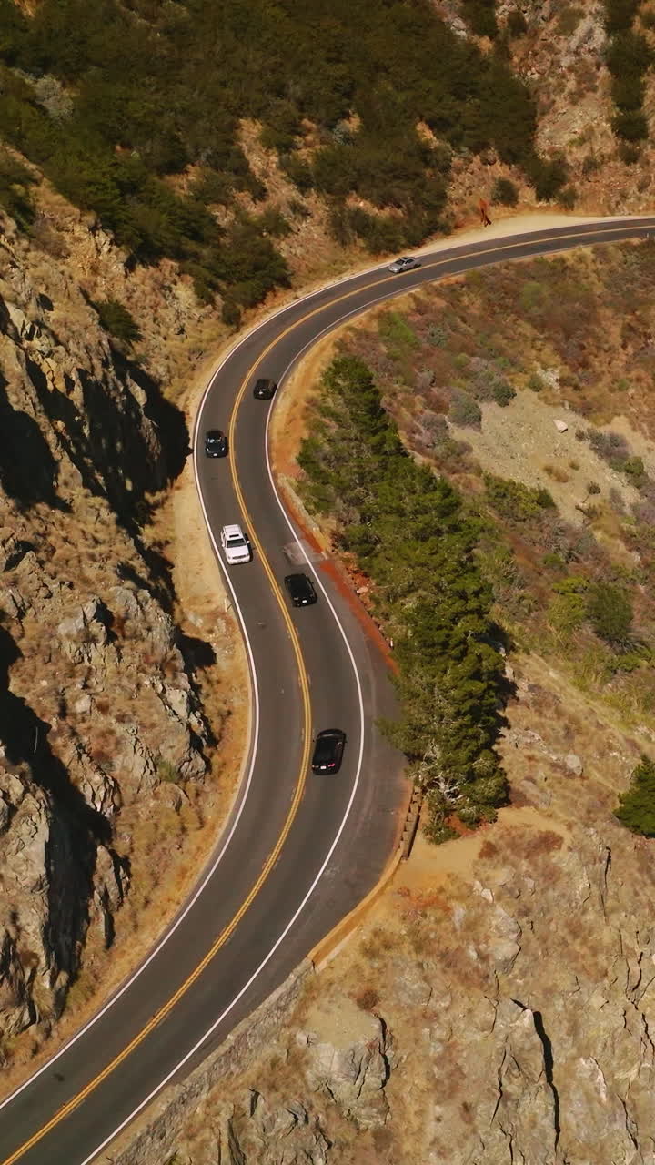 Lively road with cars moving back and forth in the mountainous landscape. Huge mountain with little greenery on sunny day from aerial view. Vertical video