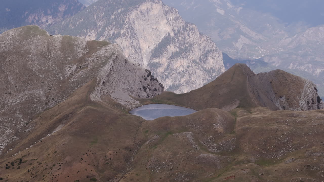 Aerial orbit approach to Dragonlake on Mount Tymfi, surrounded by rocky slopes and fog covered peaks