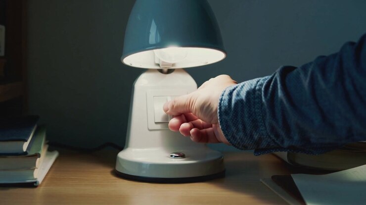 Reaching from right denim-sleeved hand toggling rocker switch on white lamp at desk, dimming light