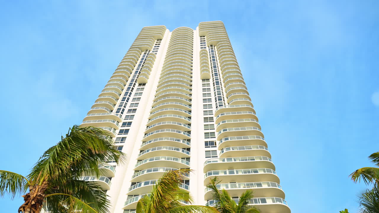 Arranha-céu branco de luxo moderno que se ergue no céu azul claro. Vista de baixo para cima de uma imponente torre residencial branca com varandas sob um céu azul brilhante em Miami Beach.