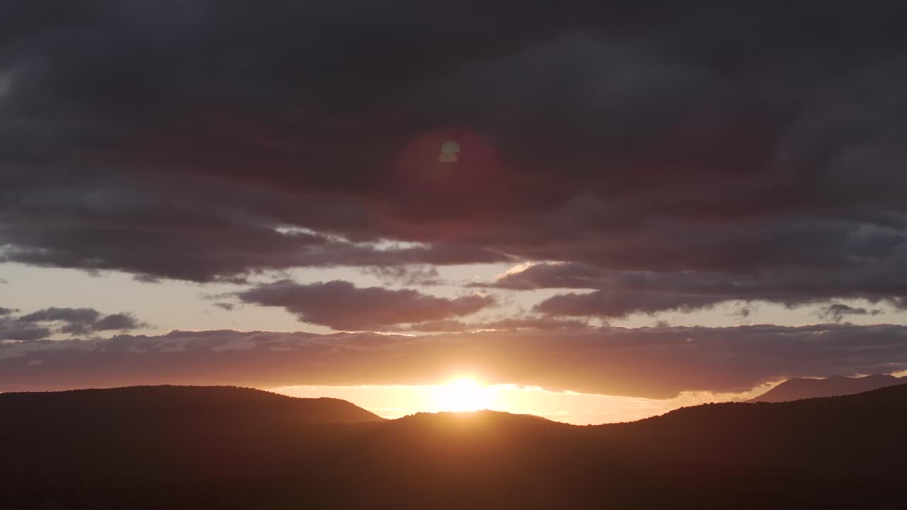 Aerial shot of a sunset in the mountains, filmed in 70mm. The dark peaks, golden rays, and a large gray cloud create a dramatic backlit scene as the drone camera slowly descends.