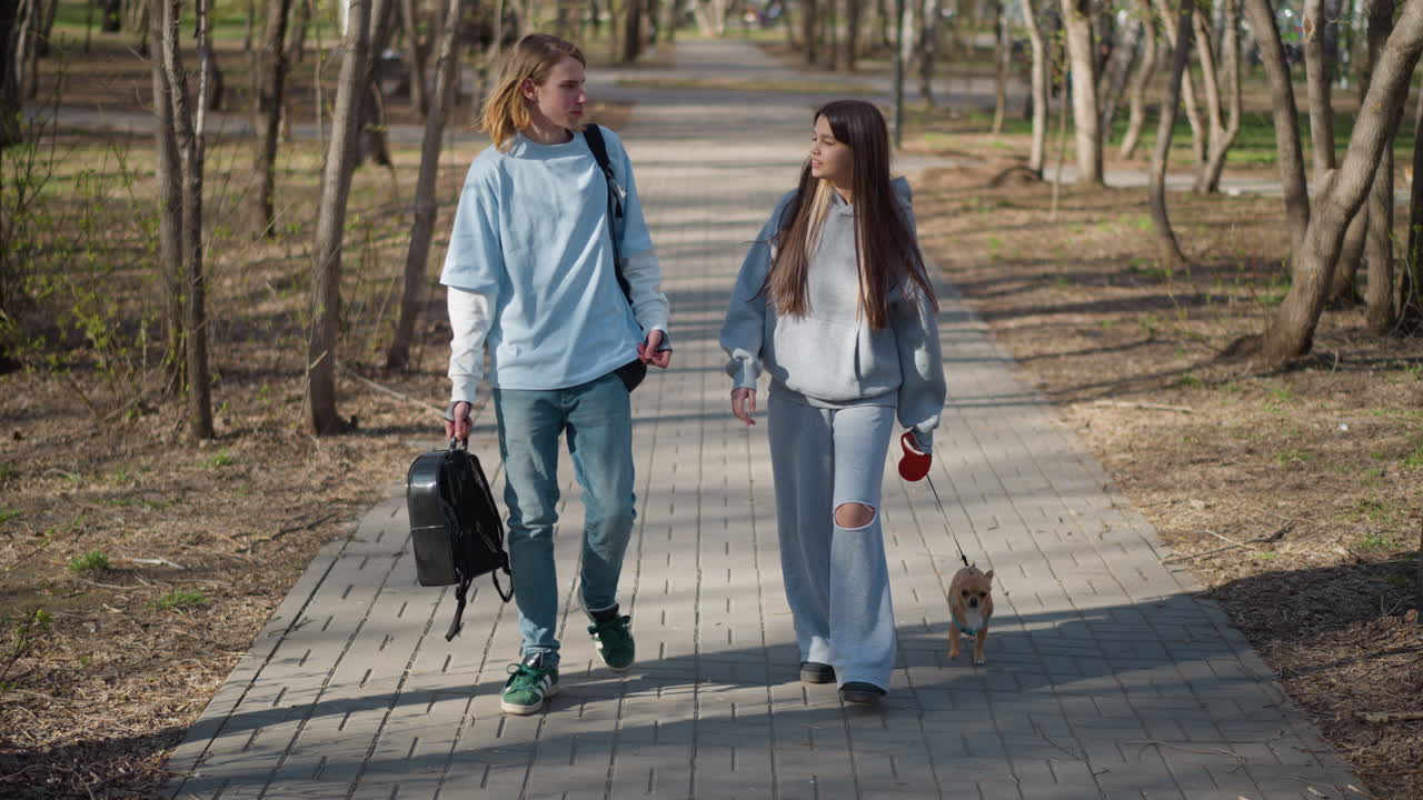 Jóvenes adultos paseando a un perro en el parque, un primer plano medio muestra una conversación animada y contacto visual mientras los amigos planean tomar un café el fin de semana, un perro pequeño a sus pies, luz cálida y un fondo frondoso