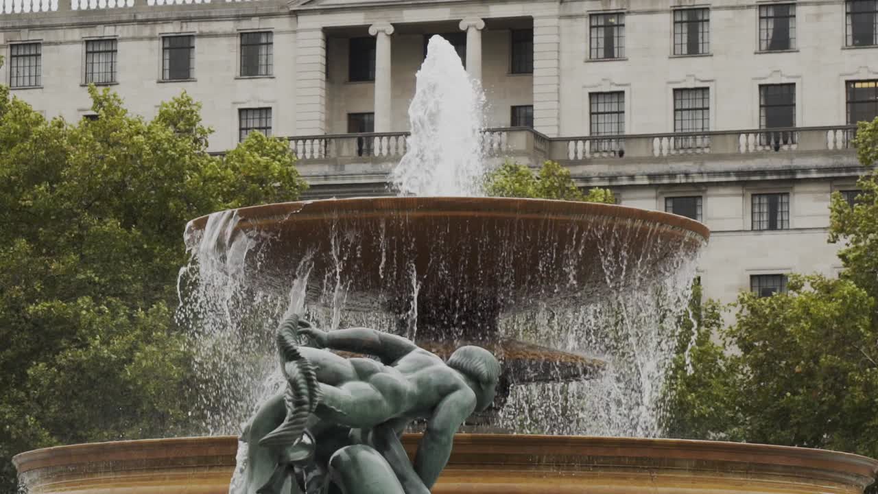 dos fuentes de agua de cemento que fluyen con una estatua de bronce verde en primer plano y un edificio de negocios urbano blanco en el fondo, londres, inglaterra, cámara lenta estática