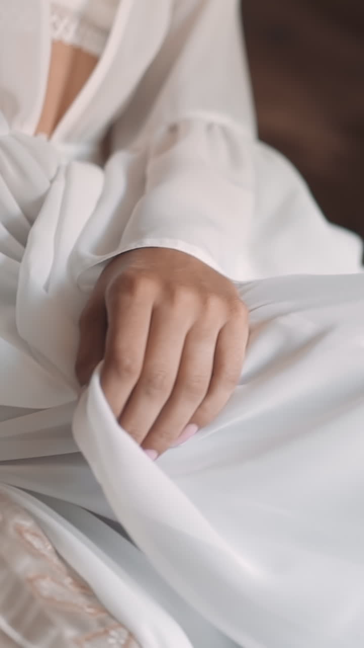 Long legged slim woman wraps up elegant white dressing gown hiding bare knees sitting on bed in hotel room closeup. Attractive woman in stylish luxury lingerie