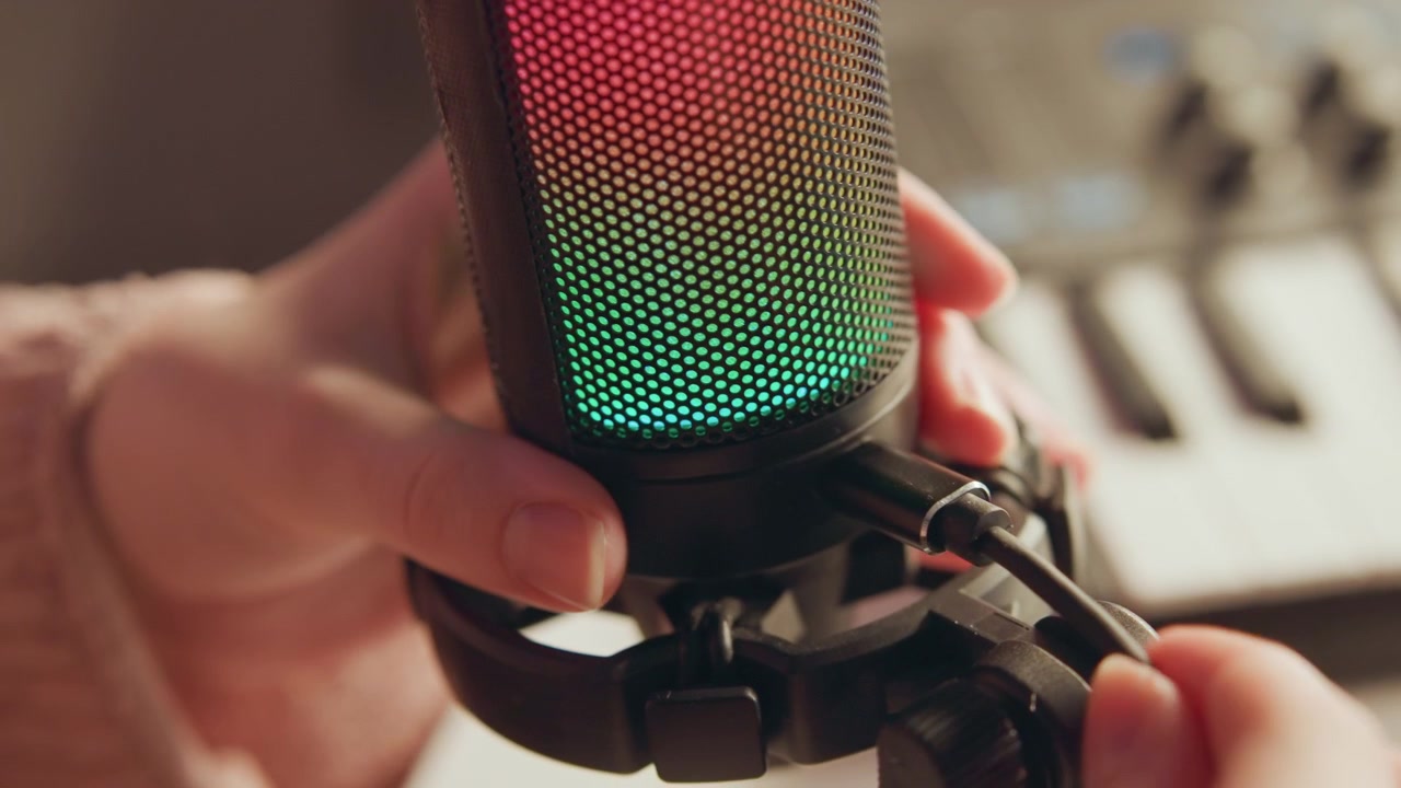 Close view of hands securing microphone inside shock mount while glowing green and red lights add color beside blurred MIDI keyboard on desk in creative home studio setup, with microphone, hand