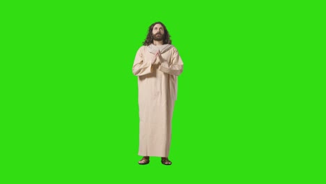 studio shot of man wearing robes and sandals with long hair and beard representing figure of jesus christ praying on green screen