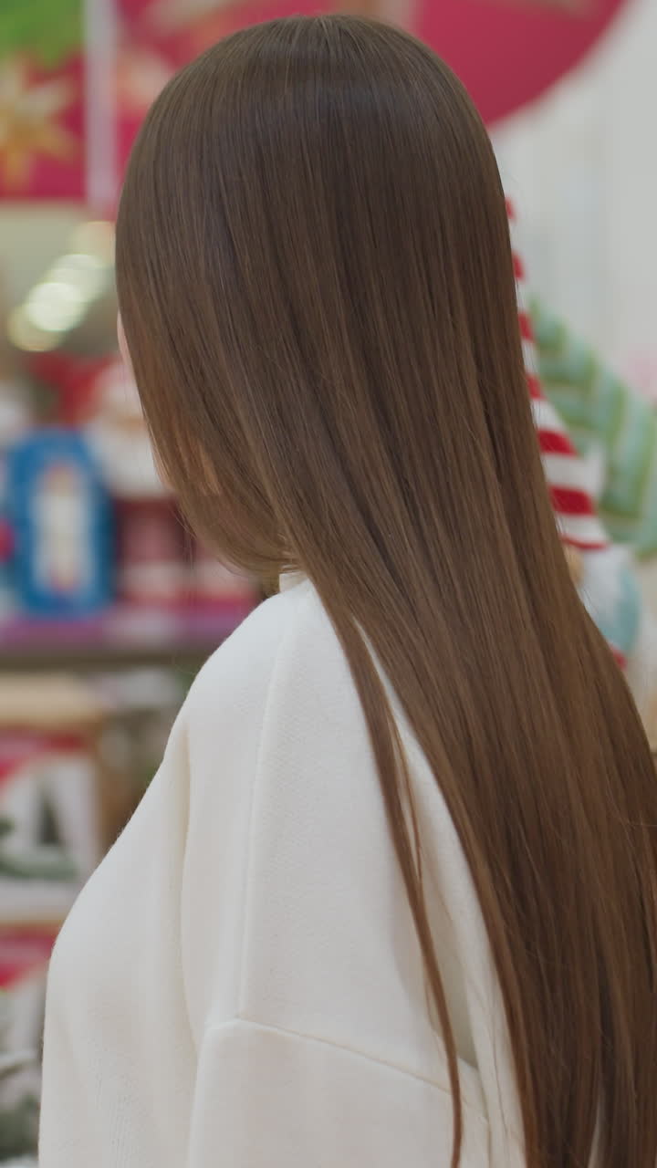 Side view of young woman walking in Christmas store filled with diverse holiday decorations and toys, she walks up to snowy themed Christmas tree as she shops for festive items