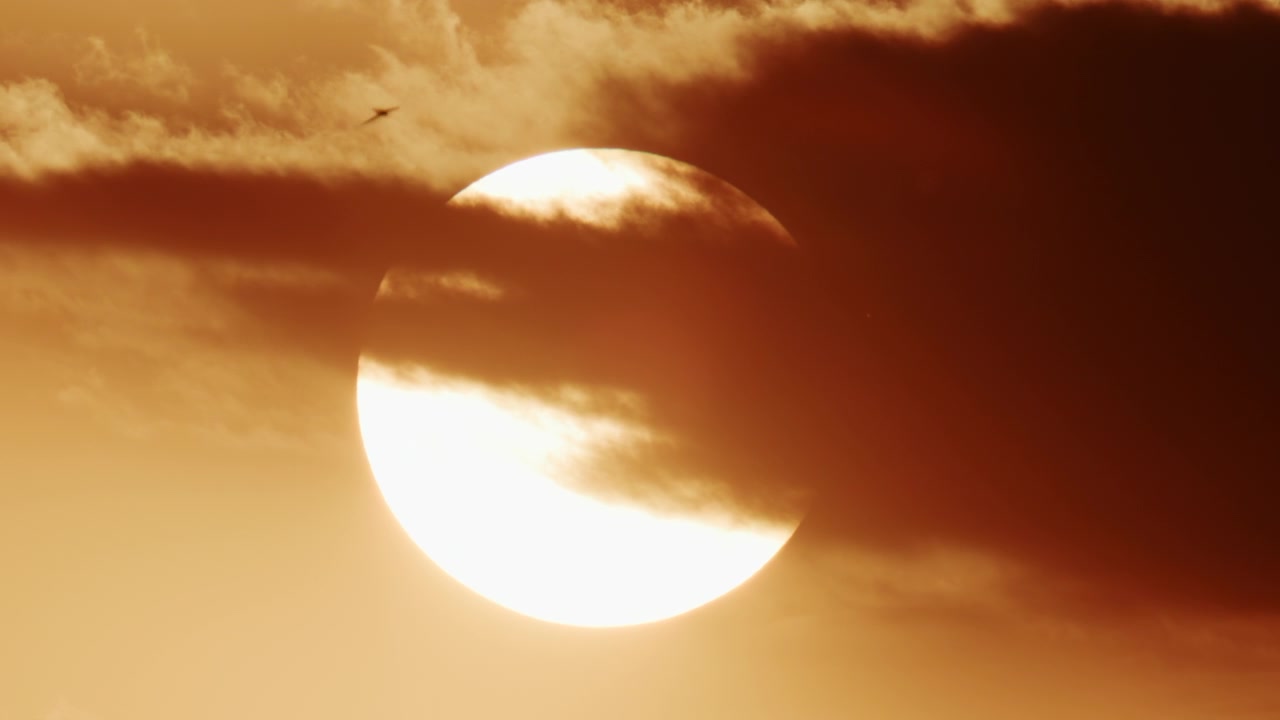 Närbild av en stark sol delvis täckt av tjocka rörliga moln, som glöder i intensiva guldorange solnedgångstoner.