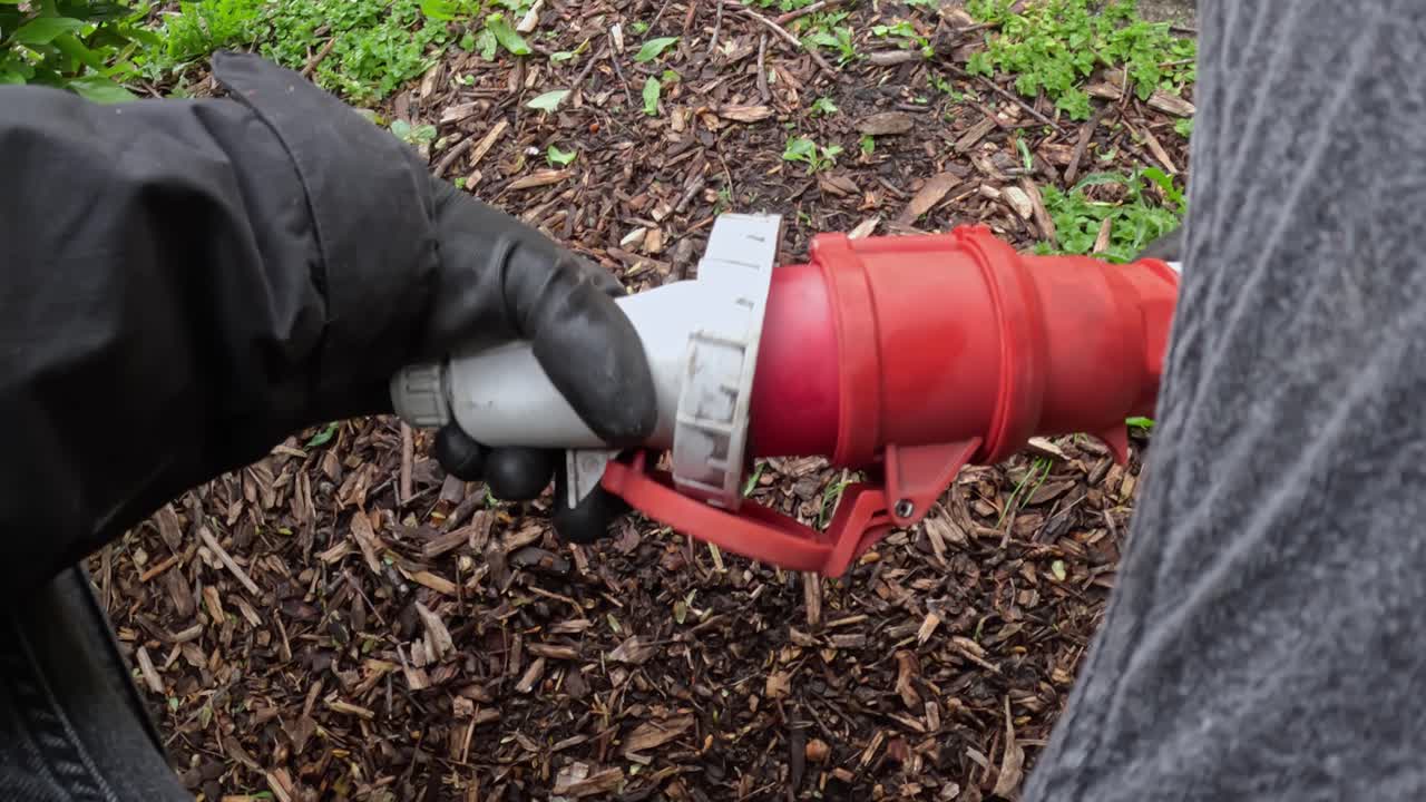 Gloved hands connect heavy-duty industrial power plug into its socket on mulch-covered soil, part of outdoor event or construction setup, red locking arm engaged, real time, close-up, first-person POV