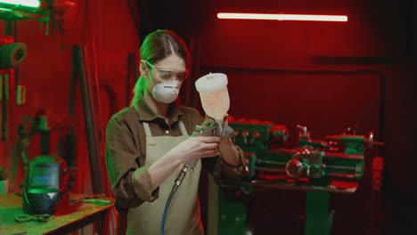 joven soldadora caucásica con gafas, delantal y máscara respiratoria trabajando en un taller de metal y revisando la máquina atomizadora para pulverización