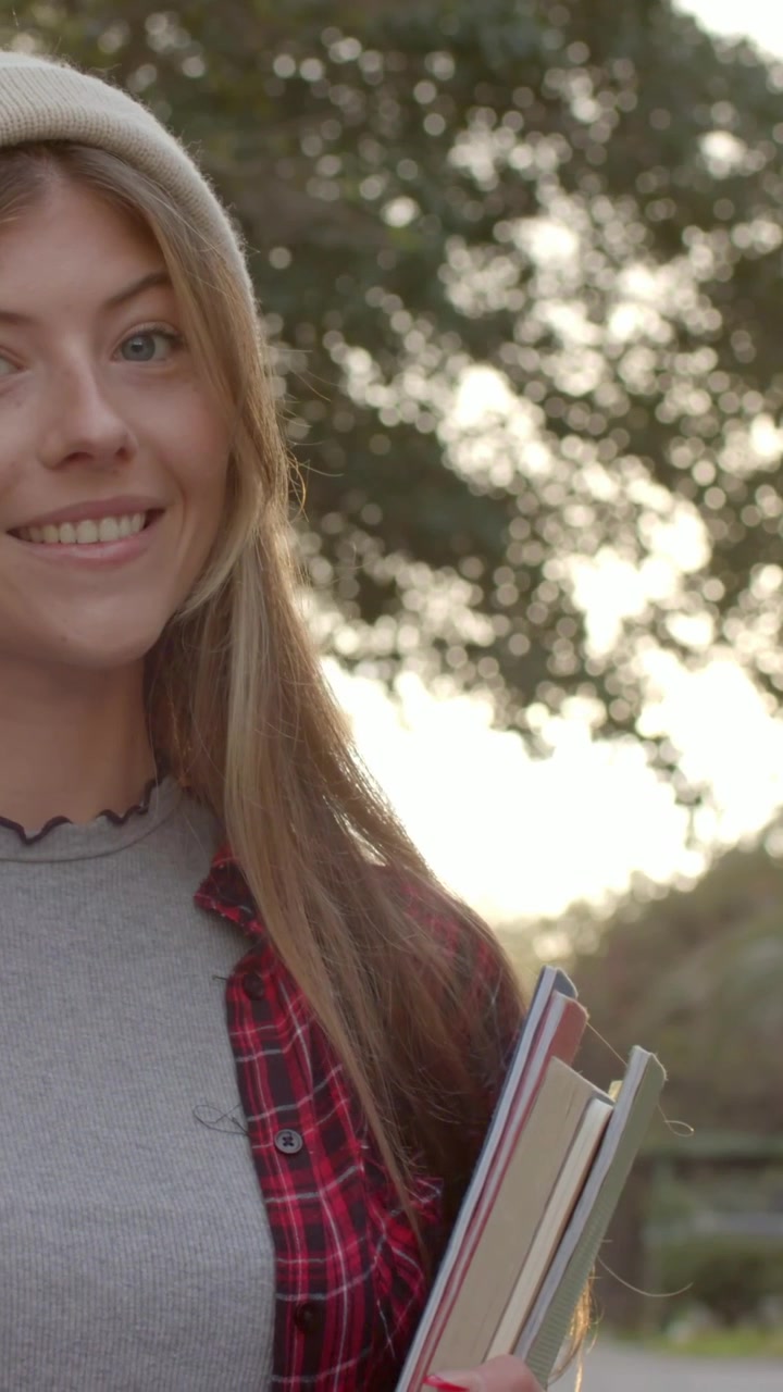 Video verticale: Donna sorridente che tiene dei libri mentre la telecamera si avvicina nel campus, indossa un berretto e una camicia a quadri.