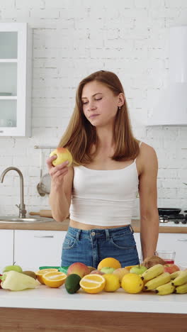 a beautiful sports girl at home in the kitchen eats a fresh apple. table with different seasonal fruits. vegetarian healthy food. diet. vertical video