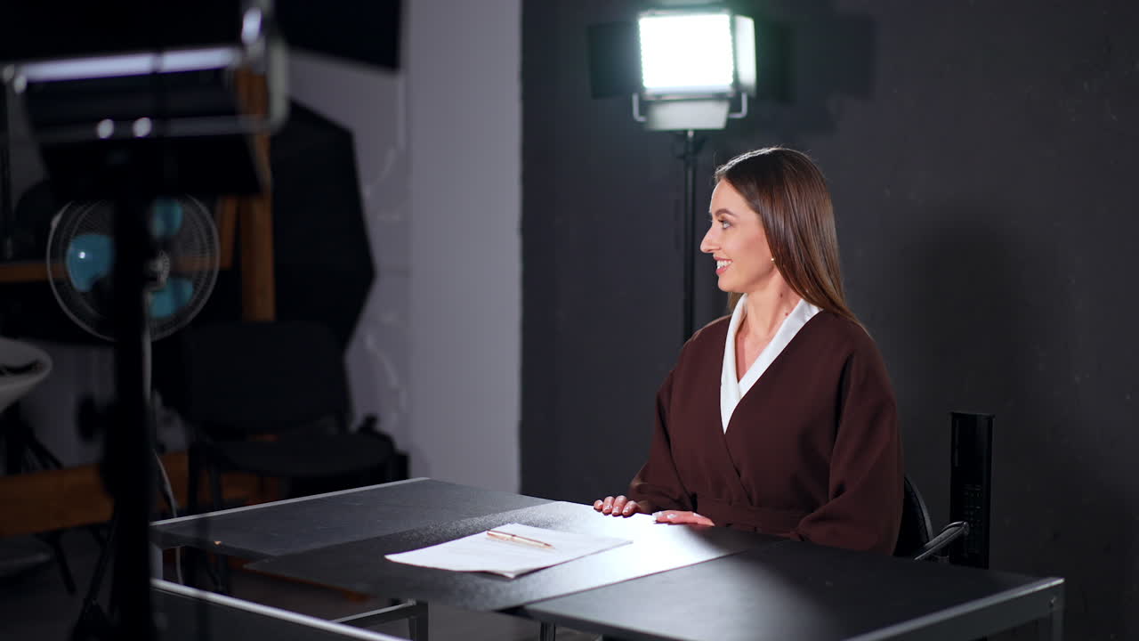 Beautiful long-haired woman in brown jacket sits down at desk. Smiling lady talks to someone backstage preparing for footage in studio.