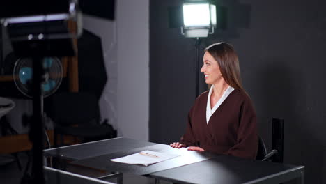 beautiful long-haired woman in brown jacket sits down at desk. smiling lady talks to someone backstage preparing for footage in studio.