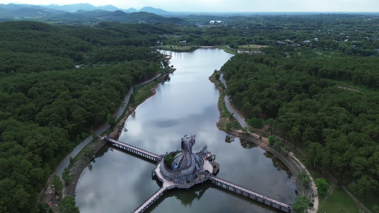 Distant semi orbit around the entire circular structure of the water park dragon statue, framed by dense forest and a large lake.