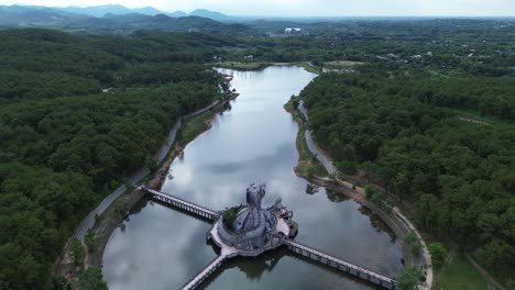 distant semi orbit around the entire circular structure of the water park dragon statue, framed by dense forest and a large lake.
