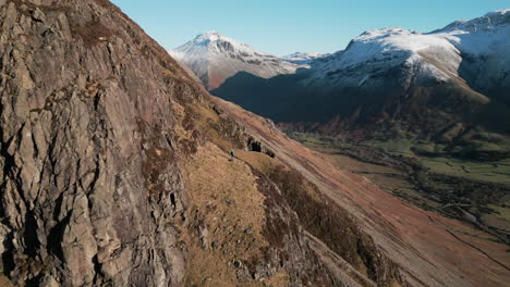 Volando-Sobre-Un-Excursionista-En-La-Ladera-De-La-Montaña-Y-Revelando-Picos-Nevados-Con-Un-Valle-Verde-En-El-Distrito-De-Los-Lagos-Wasdale,-Reino-Unido