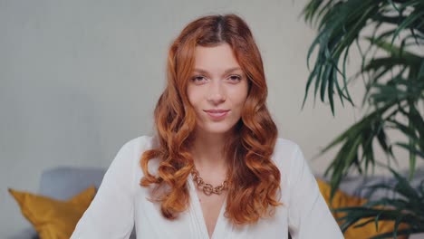 camera view redhead caucasian woman looking at camera and smiling sits in home