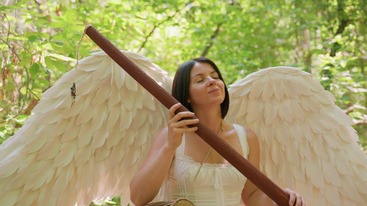 Fairy white girl with luminous wings holds wooden staff while gazing upward in forest, surrounded by sunlight and leaves, expression thoughtful and serene