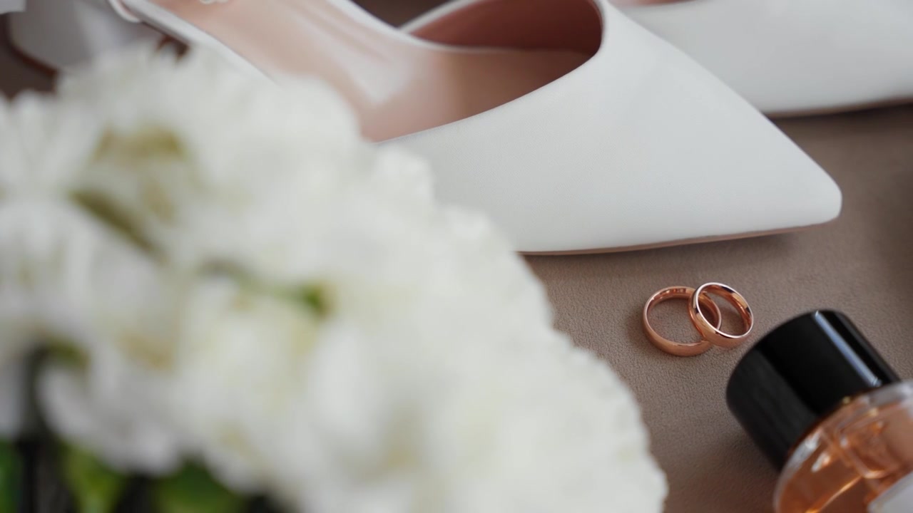 Close-up Delicado Captura Alianças de Casamento de Ouro, Sapatos Brancos Elegantes, Frasco de Perfume e Flores Brancas Suaves em Luz Natural, Perfeito para a Manhã da Noiva ou Imagens de Detalhes do Casamento