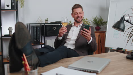 Businessman-drinking-champagne-from-glass-after-working,-putting-legs-on-table-sitting-at-office