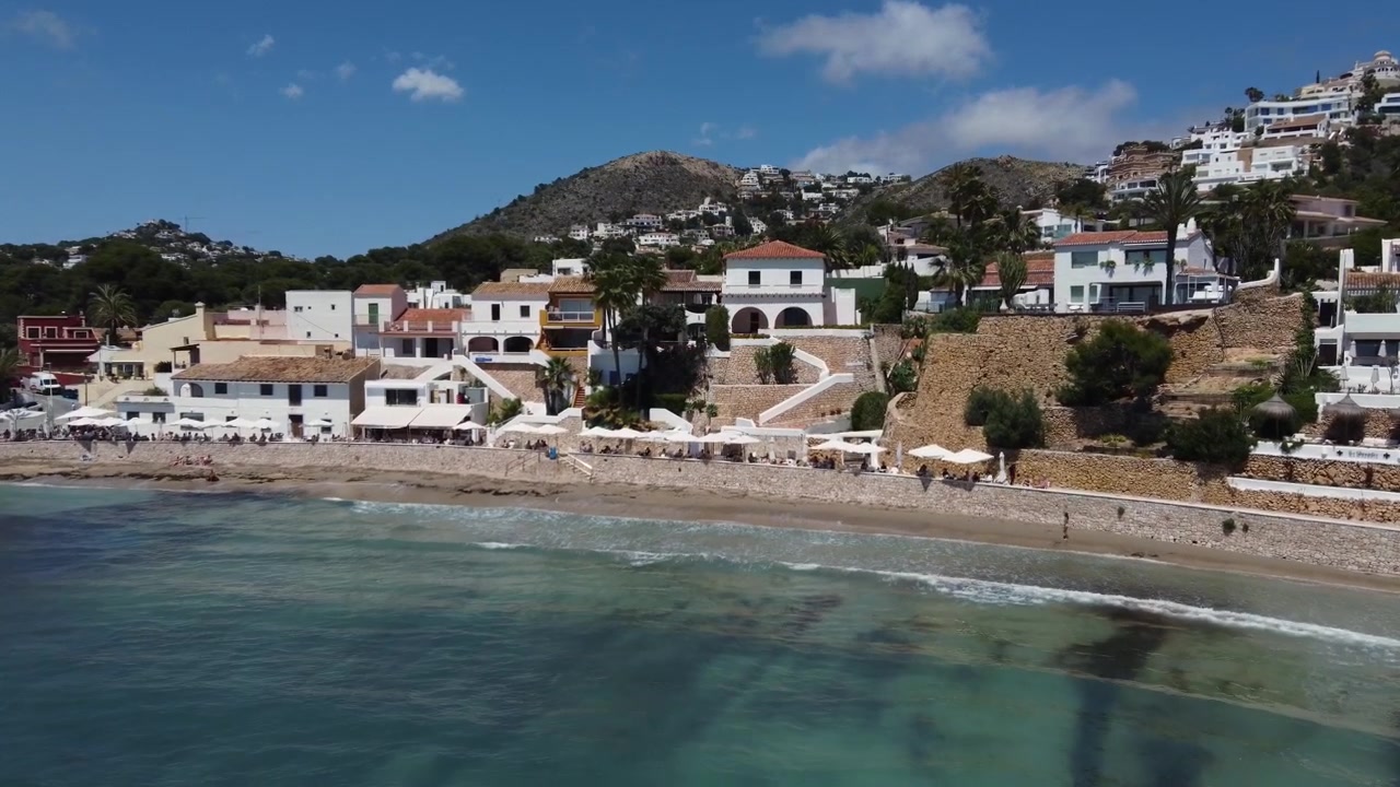 Ripresa cinematografica con drone che si avvicina alle acque turchesi e alla costa panoramica della cala di El Portet a Moraira, Spagna, mostrando uno stile di vita da spiaggia mediterraneo e soleggiato da una vista aerea.
