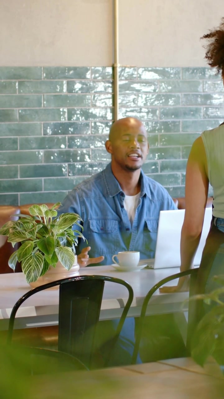 Vertical video: Approaching server in apron checking order man reaching cup by laptop at cafe