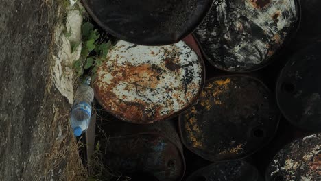 Vertical-low-close-up-panning-shot-of-old-rusty-barrels,-garbage-and-cinder-blocks-piled-causing-an-environmental-disaster