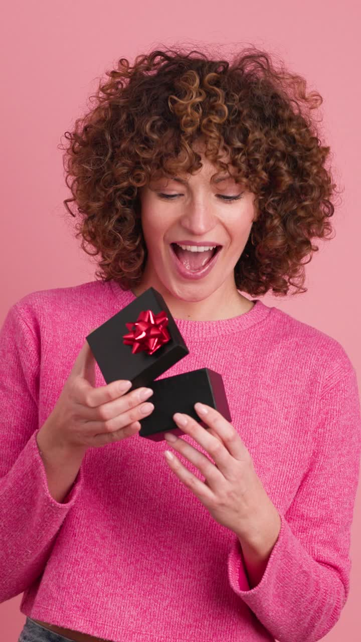 Surprised woman opening gift box in pastel pink colored studio. Vertical footage
