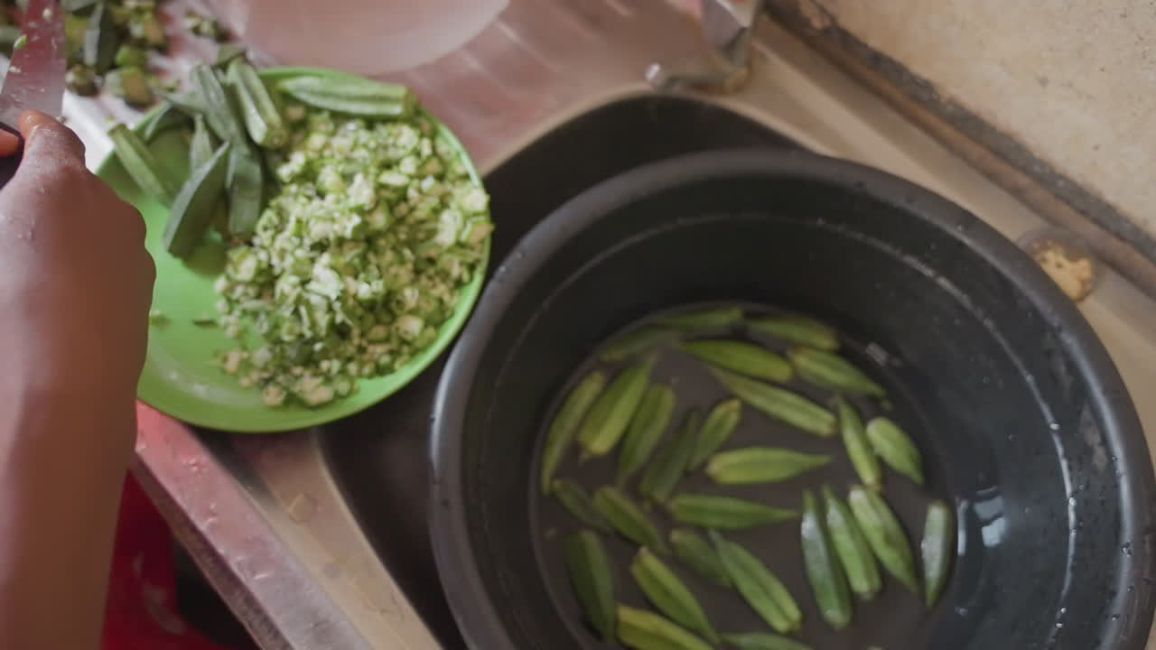 Sink hands sorting okra into pot, marketstyle prep with chopped greens on plate, large knife, wet fingertips, fastpaced communal cooking, busy countertop, closeup of motion and texture, vibrant
