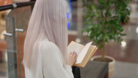 back view of youthful adult seated on bench in sweater turning pages of brown bound novel intently reading in upscale mall near potted tree during quiet reading moment with blur modern interior